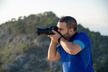 Close up of an adult latin man taking a photo with his camera at the peak of a mountain with another mountain on the background in Mallorca, Spain