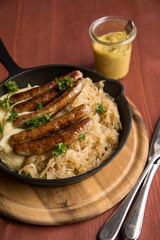 Kleine Bayrische Nürnberger Rostbratwürste mit Sauerkraut, Kartoffelpüree und Senf in rustikaler Gusseisenpfanne auf Holz Brett