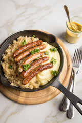 Kleine Bayrische Nürnberger Rostbratwürste mit Sauerkraut, Kartoffelpüree und Senf in rustikaler Gusseisenpfanne auf Holz Brett und Marmor Tisch