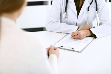 Fototapeta premium Woman doctor and patient sitting and talking at medical examination at hospital office, close-up. Therapist filling up medication history records. Medicine and healthcare concept