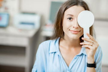 Young woman checking vision covering one eye with an ophthalmic scapula during a medical examination at the ophthalmological office - obrazy, fototapety, plakaty