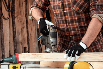 A man carpenter cuts a wooden beam using an electric jigsaw, male hands with an electric jigsaw...