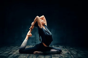 Wall murals Yoga school Young woman practicing yoga doing one legged king pigeon pose in dark room