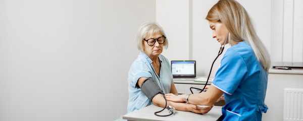 Nurse measuring blood pressure of a senior woman patient during an examination in the clinic. Senior care concept. Wide view with copy space - obrazy, fototapety, plakaty