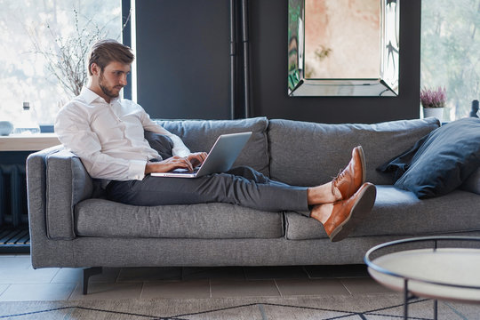 Multitasking. Handsome Young Man Working With Laptop While Sitting On The Couch In Office