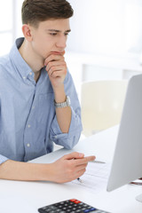 Fototapeta premium Business man sitting and working with computer and calculator in white office. Student guy studying at home or young accountant checking balance. Tax, audit and bookkeeping concept