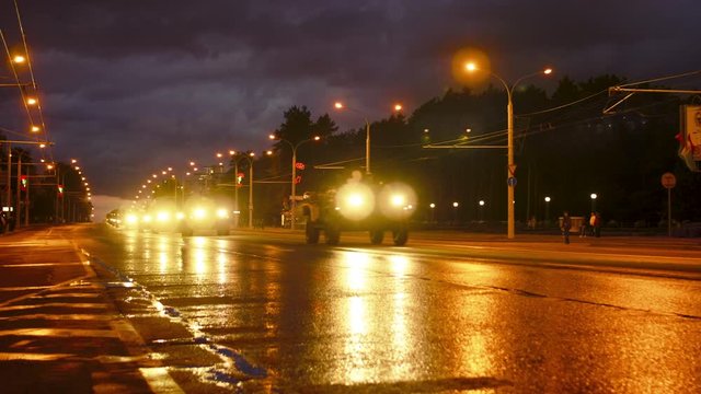 A convoy of military equipment rides through the city at night with headlights on