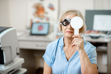 Senior woman checking vision with eye test glasses and scapula during a medical examination at the ophthalmological office - obrazy, fototapety, plakaty