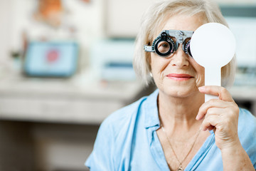 Senior woman checking vision with eye test glasses and scapula during a medical examination at the ophthalmological office - obrazy, fototapety, plakaty
