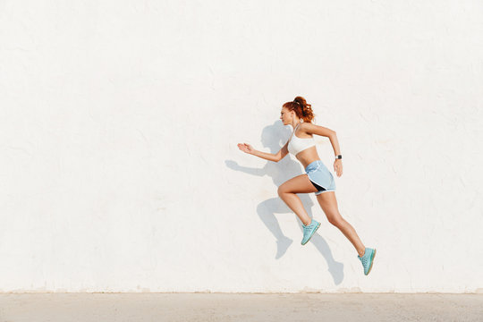 Image of redhead young woman running along white wall while doing workout in morning