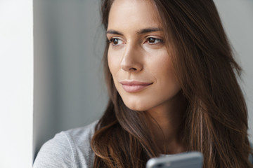 Close up of a young beautiful girl standing leaning on a gray wall over gray background, using mobile phone