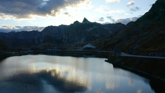 Aerial climb over Grand Saint Bernard alpine pass at sunset with clouds reflecting in the lake. Border between Italy and Switzerland