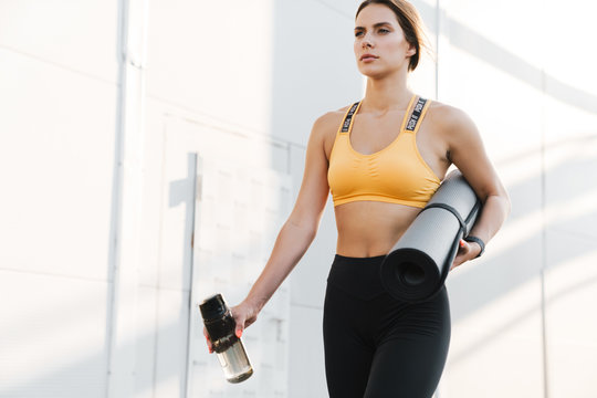 Image of lovely woman walking with yoga mat and water bottle outdoors