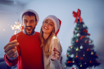 Happy caucasian couple with santa hats on heads holding sprinklers while hugging and standing in living room. In background is christmas tree. Selective focus on sprinklers.