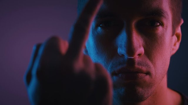 Portrait of Man Showing Fuck You in neon lights. Aggressively tuned male shows middle finger in a dark room lit by neon light.