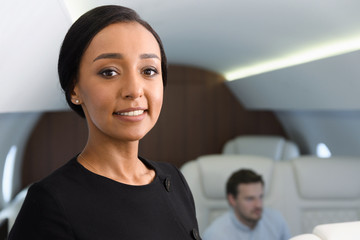 Stewardess portrait in private jet. Female biracial flight attendant smiling inside of business airplane cabin with passengers on background.
