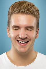 Fototapeta premium Caucasian young man's close up portrait on blue studio background. Beautiful male model with well-kept skin. Concept of human emotions, facial expression, sales, ad, beauty. Smiling, laughting.