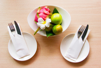 Knives and forks wrapped in napkins lie on white plates. Nearby is a vase of exotic fruits and a flower plumeria. The view from the top. The concept of the table setting.