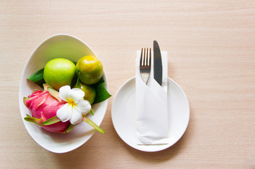 Knife and fork wrapped in a napkin lying on a white plate. Nearby is a vase of exotic fruits and a flower plumeria. The view from the top. The concept of the table setting.