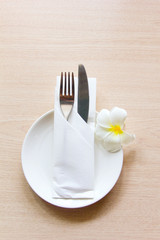 Knife and fork wrapped in a napkin lying on a white plate decorated with a flower plumeria. The view from the top. The concept of the table setting.
