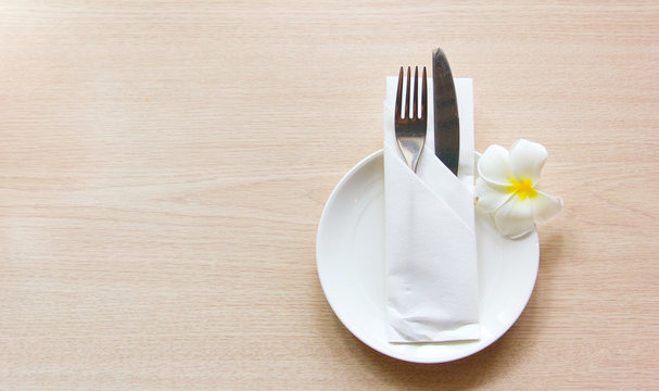 Knife and fork wrapped in a napkin lying on a white plate decorated with a flower plumeria. The view from the top. The concept of the table setting.