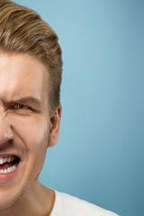Fototapeta premium Caucasian young man's close up portrait on blue studio background. Beautiful male model with well-kept skin. Concept of human emotions, facial expression, sales, ad, beauty. Angry screaming.