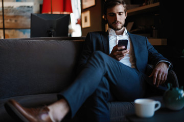 Naklejka na ściany i meble Smiling bearded man making text message on smartphone while sitting sofa at modern office.
