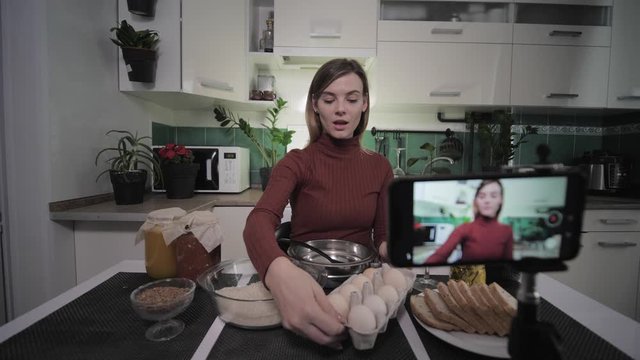 woman blogger, sitting at a table with groceries in the kitchen writes on smartphone for her subscribers a video recipe, raises a glass of wine and drinks while making a toast