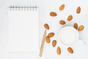 Almond milk in cup, notebook, pencil, nuts on white concrete background. Non-dairy, vegan, recipe concept. Top view, flat lay, copy space, mock up