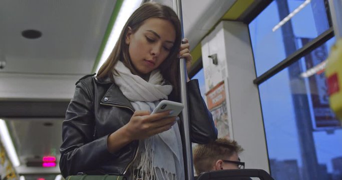 Caucasian young atractive and stylish woman standing in the tram and holding a handrail while tapping and typing on the phone in the evening.