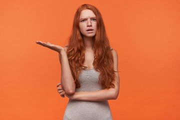 Studio shot of pretty young foxy haired female in grey shirt looking at camera with confused face, frowning embarrassed and raising palm up, isolated over orange background