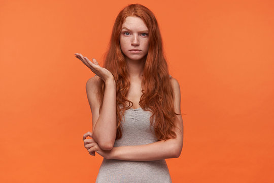 Attractive Young Red Haired Female With Wavy Lond Hair Posing Over Orange Background In Casual Clothes, Looking At Camera With Raised Eyebrow And Raising Palm Perplexedly