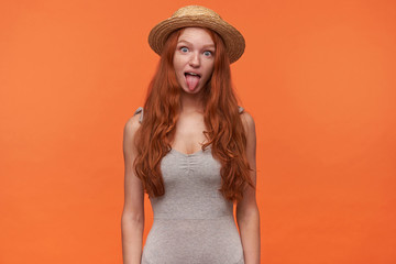 Funny indoor shot of joyful pretty young redhead woman with long hair, looking to camera and showing tongue, posing over orange background in casual clothes