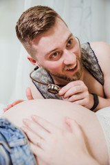 A young man with enthusiasm, joy and surprise listens to the naked belly of his beautiful pregnant wife with a stethoscope.