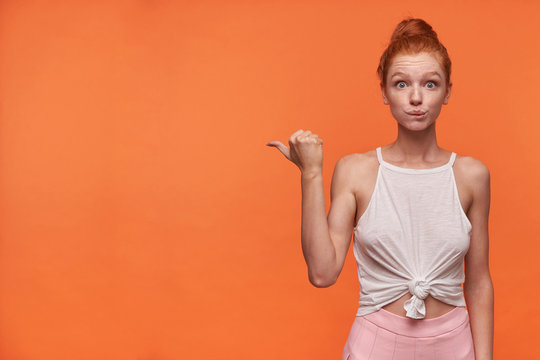 Indoor photo of lovely young redhead woman with bun hairstyle wearing white top and pink skirt, standing over orange background and pointing aside with thumb, wrinkling forehead with raised eyebrows