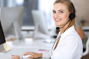 Blonde business woman using headset for communication and consulting people at customer service office. Call center. Group of operators at work at the background