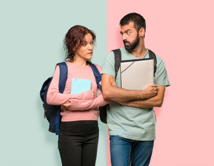 Two students with backpacks and books having doubts and with confuse face expression while bites lip on colorful wall