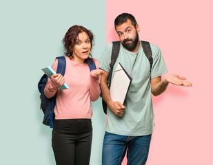 Two students with backpacks and books having doubts and with confuse face expression on colorful wall