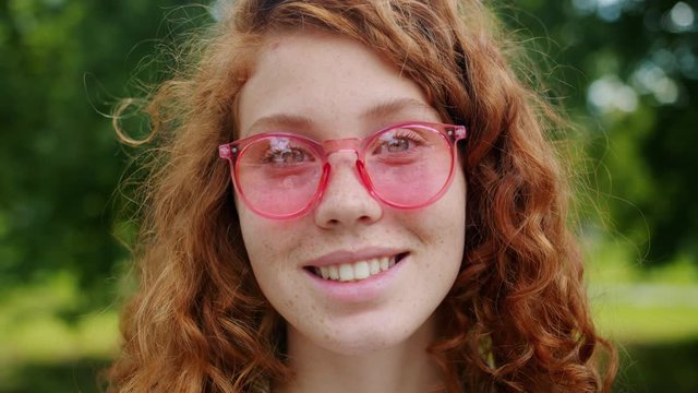 Close-up portrait of pretty redhead girl with freckles wearing sunglasses in park on summer day smiling looking at camera. Beautiful people and nature concept.