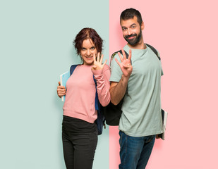 Two students with backpacks and books happy and counting four with fingers on colorful wall