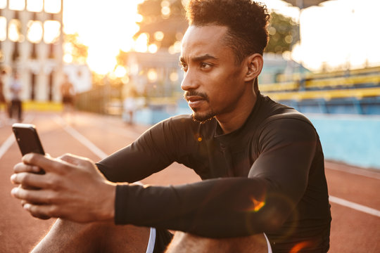Image of young african american man holding smartphone at sports ground