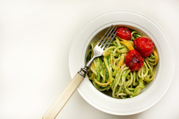 Italian Pasta with zucchini noodles with Avocado Sauce pesto and roasted tomato in white plate and fork. Top view white background. Healthy food concept.