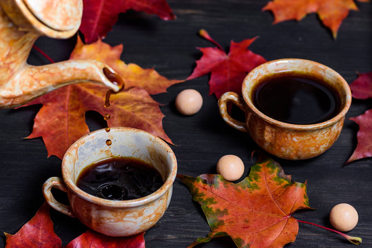 Morning coffee in mugs and a coffee pot pouring coffee into a mug on a dark wooden background with maple leaves and scattered sweets