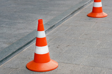 Two road cones on a square gray tile. Orange cones with white stripes. Fencing during road...