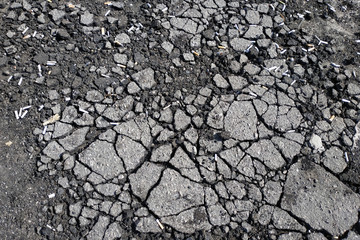 Cracked asphalt pavement with cigarette butts scattered on it. Cigarette butts on the destroyed asphalt near the entrance. Problems due to poor quality road surface in the city.
