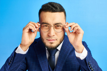 Portrait of Kazakh Asian businessman in a suit and spectacles, looking curiously at you, squinting, trying to look closer on a blue background in the studio