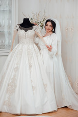 Wedding. Bride in beautiful robe near the mannequin with dress indoors at home. Trendy wedding style shot. Sexy bride. Bride touch  your white wedding dress with pearl nails. Fashion bride