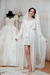 Fototapeta premium Bride's preparation before the wedding ceremony. Bride in beautiful robe near the mannequin with dress indoors at home. Fashion bride. Bride with hair style and fashion make up. Wedding. 