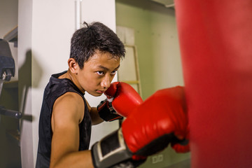 young handsome and fierce teenager boy doing fight workout punching heavy bag looking cool and badass at fitness club training hard using gloves