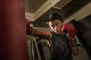 young handsome and fierce teenager boy doing fight workout punching heavy bag looking cool and badass at fitness club training hard using gloves